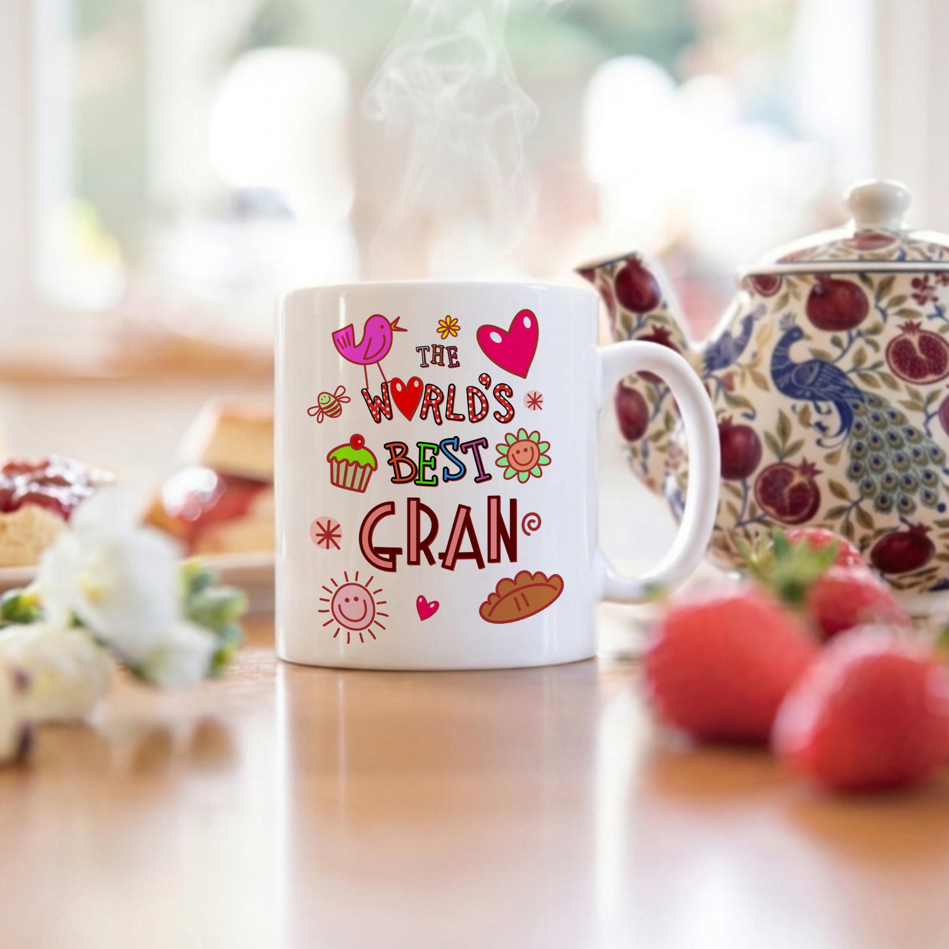 White mug with colorful text 'The World's Best Gran' on a table with a teapot and fruit in the background.
