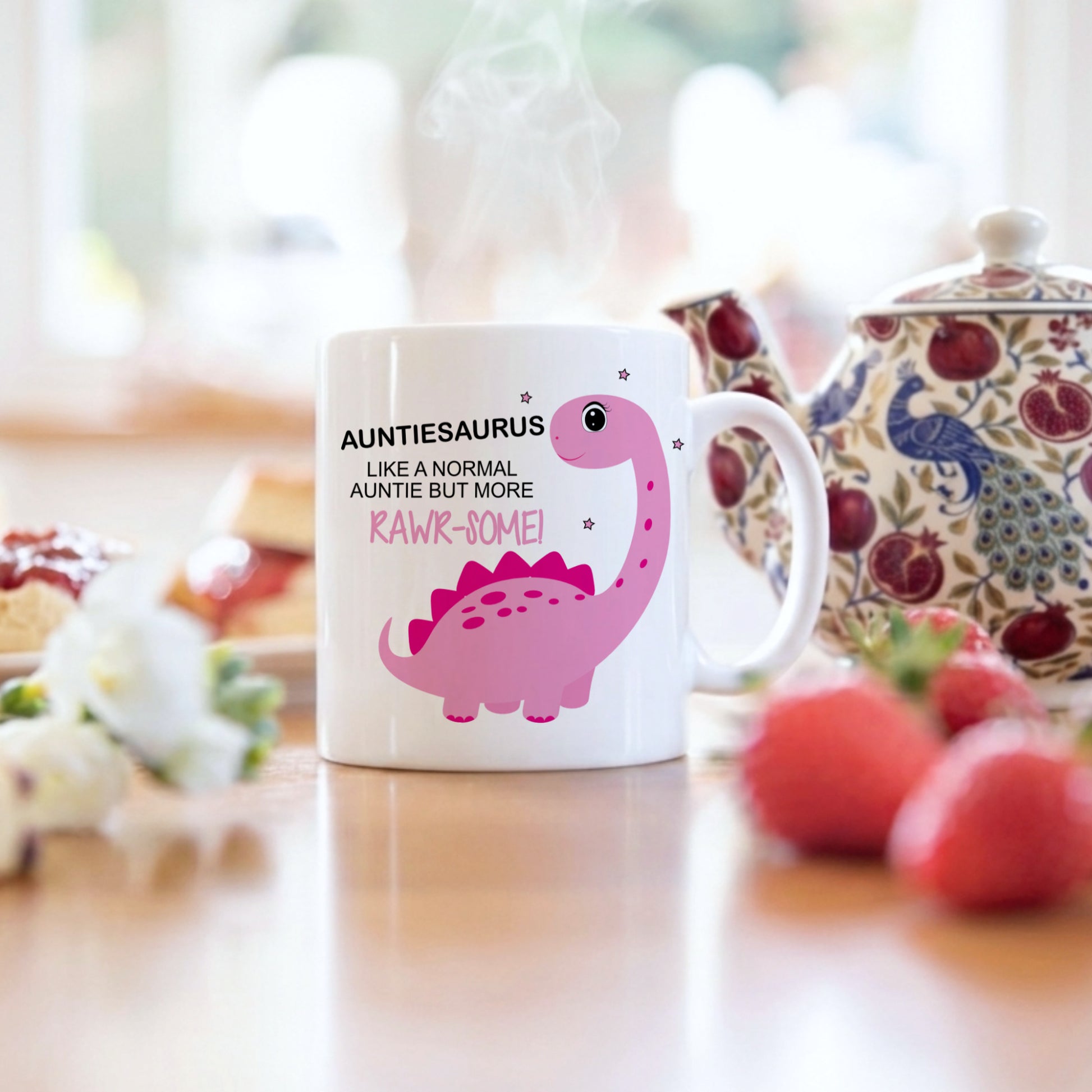 A white ceramic mug with a pink dinosaur graphic and the text 'AUNTIESAURUS LIKE A NORMAL AUNTIE BUT MORE RAWR-SOME!' sits on a wooden table in a kitchen setting, with a teapot, strawberries, and flowers blurred in the background.