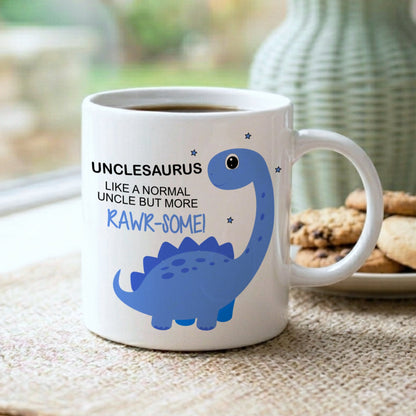 A white ceramic coffee mug with a blue dinosaur graphic and the text 'UNCLESAURUS LIKE A NORMAL UNCLE BUT MORE RAWR-SOME!' sits on a woven placemat in a kitchen setting, with cookies on a plate and a green vase blurred in the background.