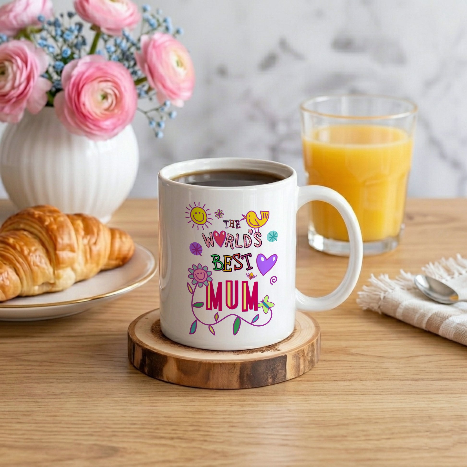 White mug with 'The World's Best Mum' text on a wooden coaster, surrounded by coffee, croissant, and juice.