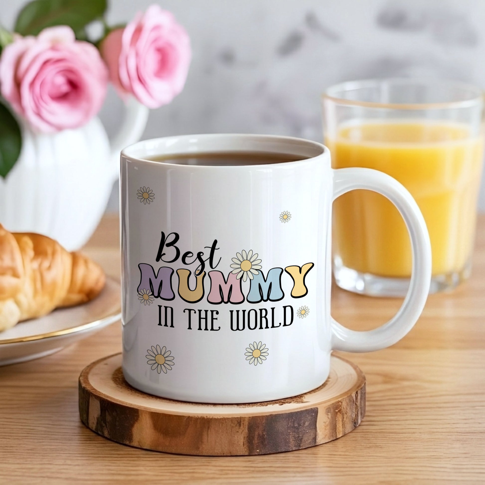White mug with 'Best Mummy in the World' text on a wooden coaster, surrounded by coffee, juice, and pastries.