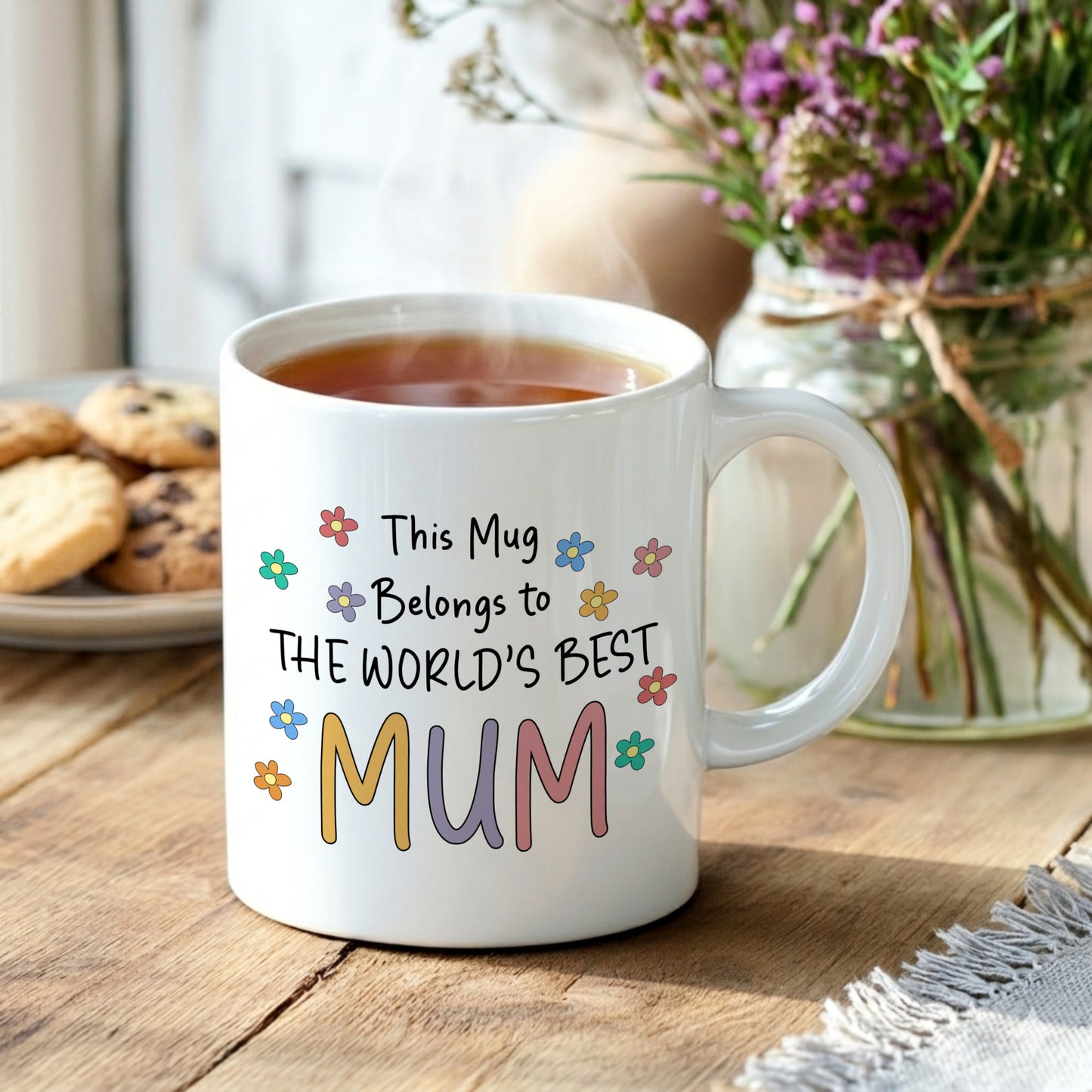 White mug with colorful text 'This Mug Belongs to THE WORLD'S BEST MUM' on a wooden table with tea and cookies.