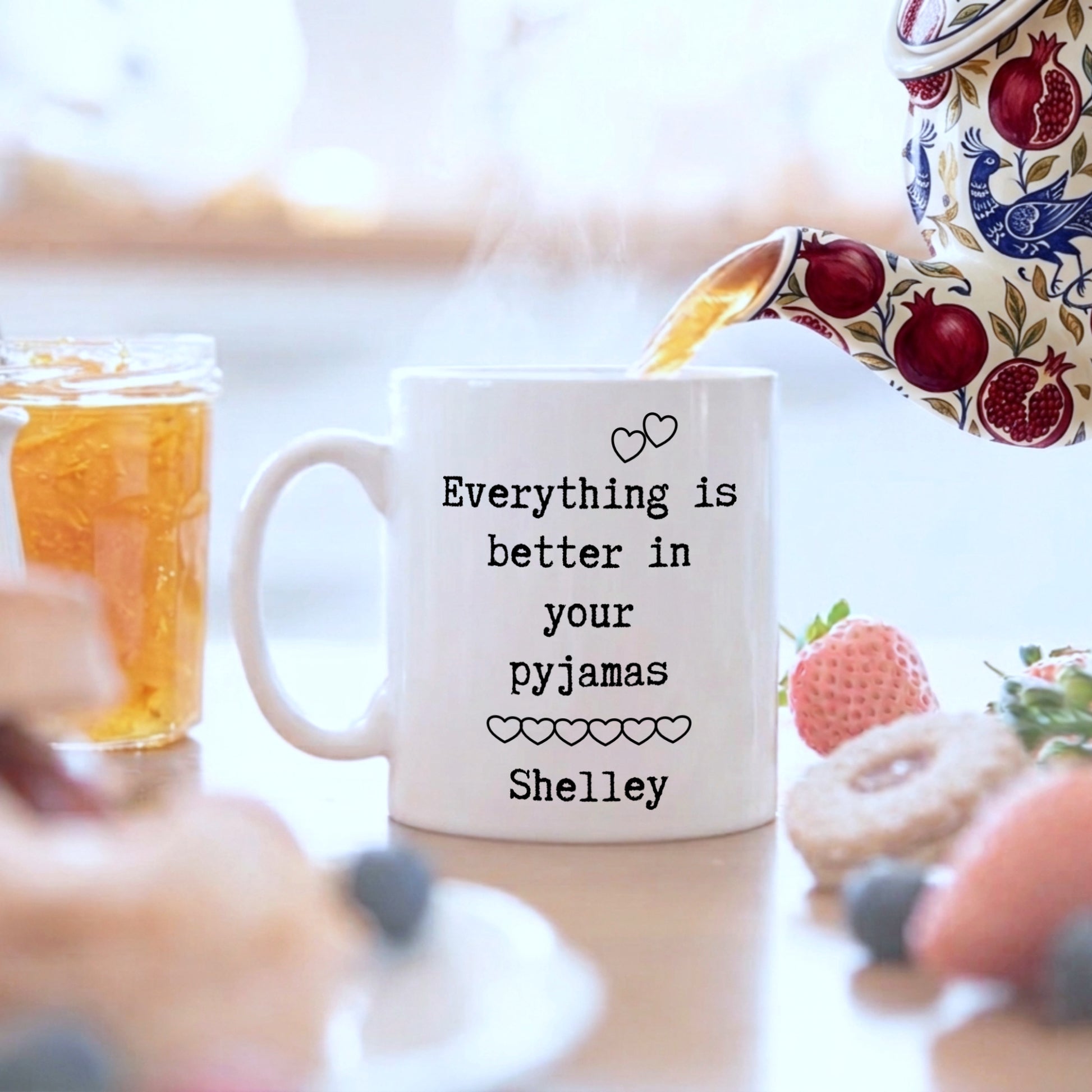 A white ceramic mug with black text reading 'Everything is better in your pyjamas Shelley' sits on a table with a teapot pouring tea into it, surrounded by breakfast items like a strawberry, a donut, blueberries, and a jar of honey.