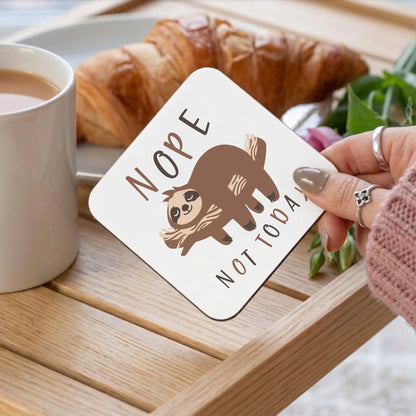 'Nope Not Today' sloth coaster held by a woman with grey nails, displayed on a wooden tray alongside a cup of tea and a croissant