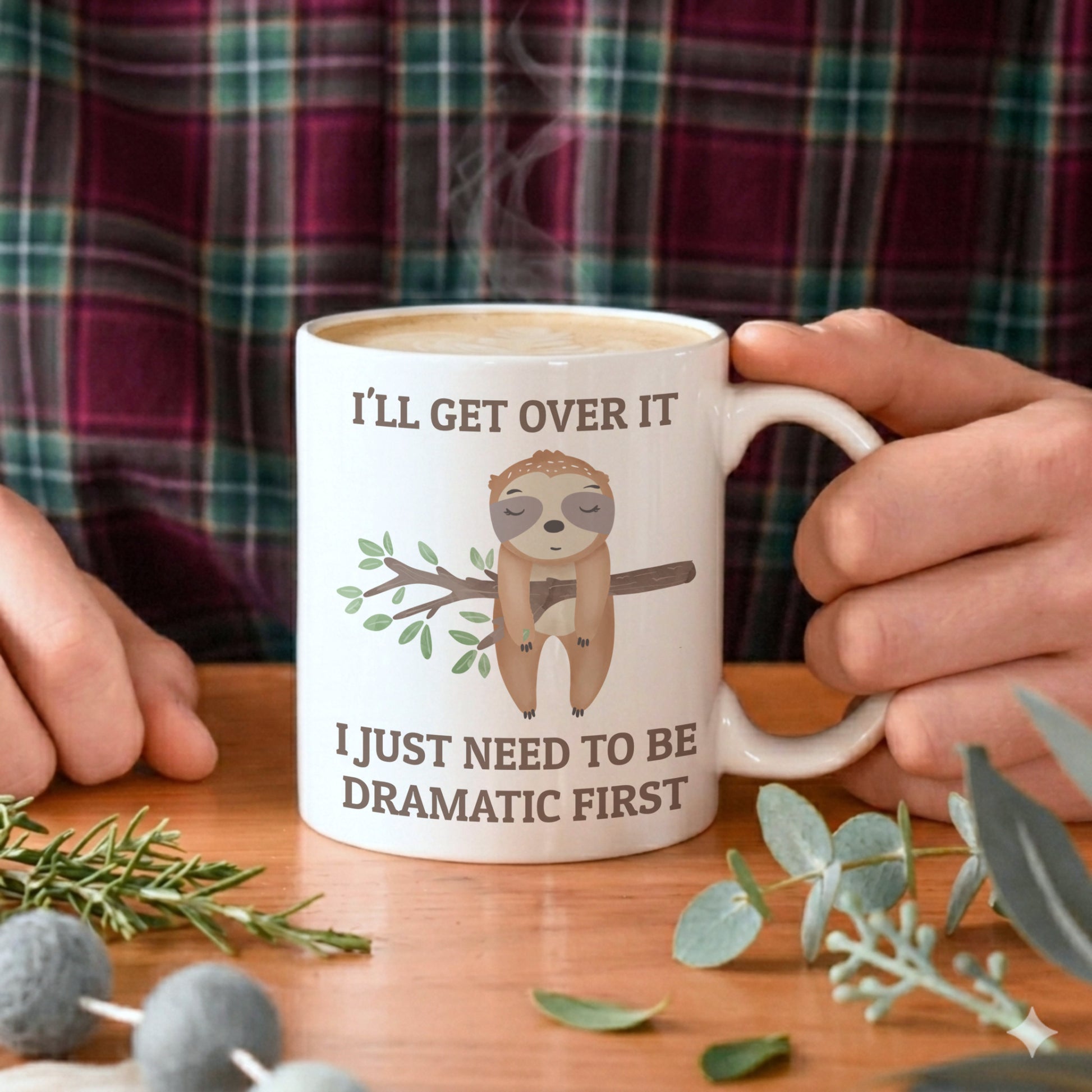 Person holding a mug with a sloth graphic and humorous text on a wooden table.