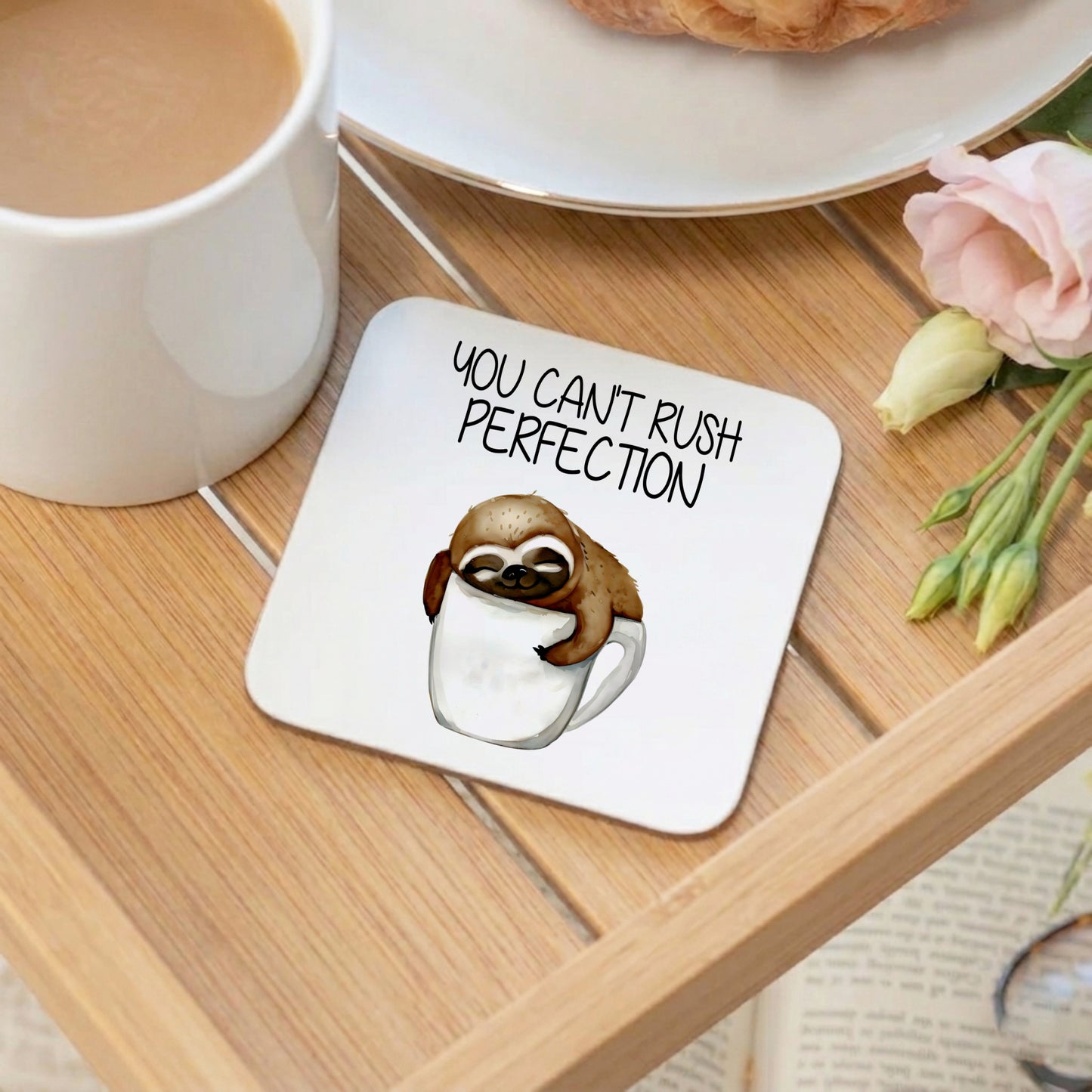 Coaster with sloth illustration and 'You can't rush perfection' text on a wooden table with coffee and flowers.
