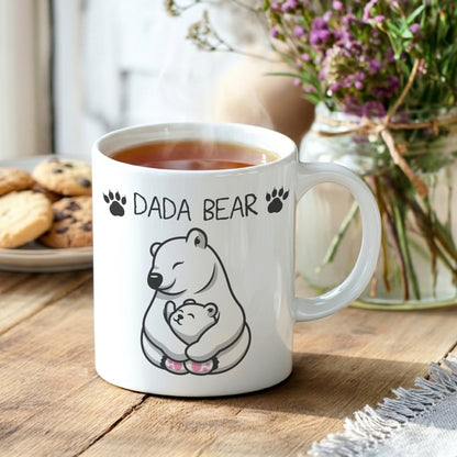 A white ceramic mug filled with tea sits on a wooden table, printed with the words 'DADA BEAR' and a black-and-white polar bear illustration holding a small cub. The mug is styled in a cozy indoor setting with cookies and a vase of flowers in the background.