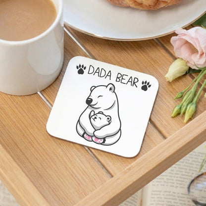 A square white coaster with rounded corners sits on a wooden table, printed with a black-and-white polar bear hugging a smaller bear and the words “DADA BEAR” with paw prints. A white mug, a plate with a pastry, and pink flowers are partly visible around it.