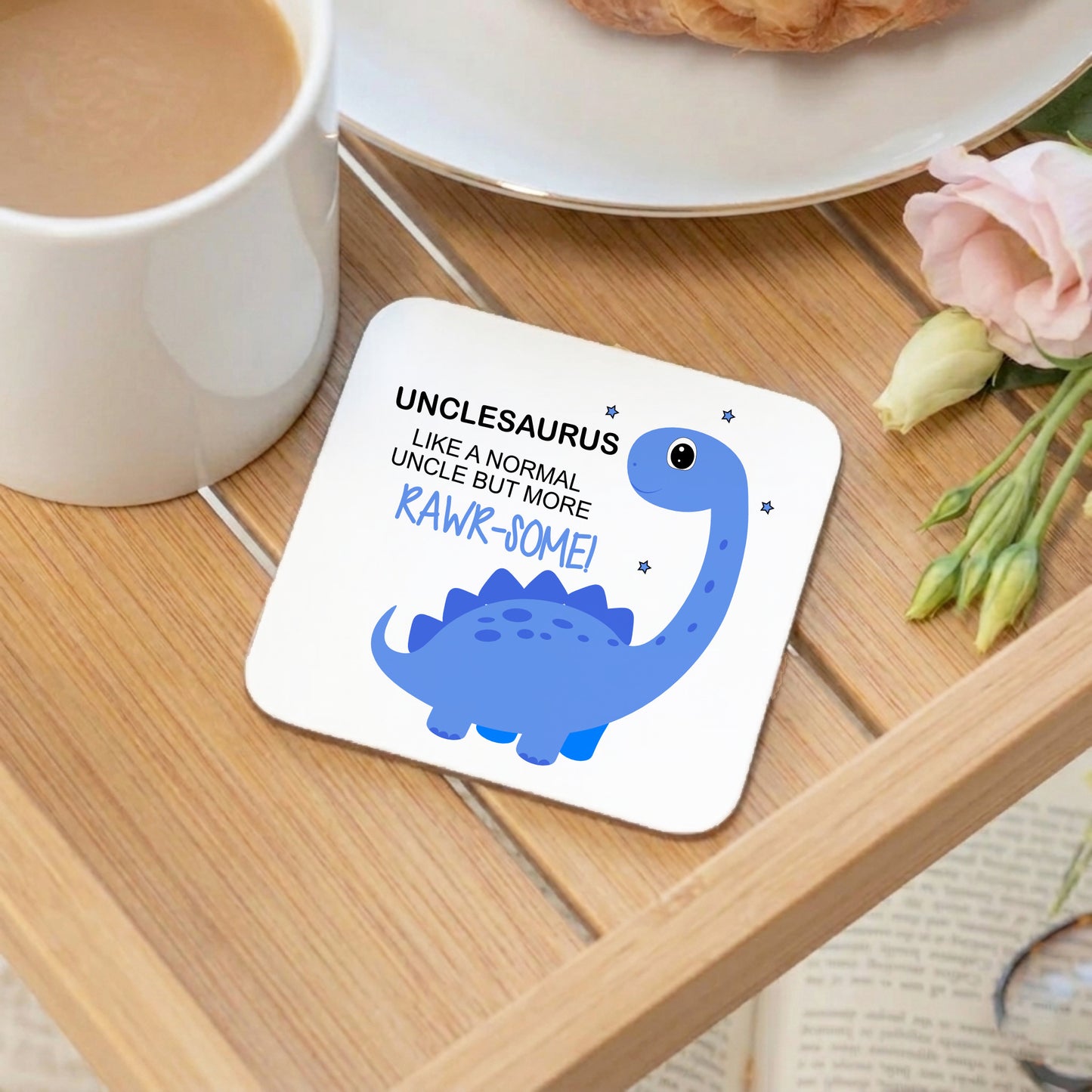 A square white coaster with rounded corners sits on a wooden tray, printed with a blue dinosaur illustration and the text 'UNCLESAURUS LIKE A NORMAL UNCLE BUT MORE RAWR-SOME!'. A white mug, plate, pastry, and pink flower are visible around it in a styled setting.