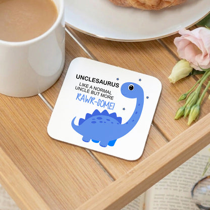 A square white coaster with rounded corners sits on a wooden tray, printed with a blue dinosaur illustration and the text 'UNCLESAURUS LIKE A NORMAL UNCLE BUT MORE RAWR-SOME!'. A white mug, plate, pastry, and pink flower are visible around it in a styled setting.