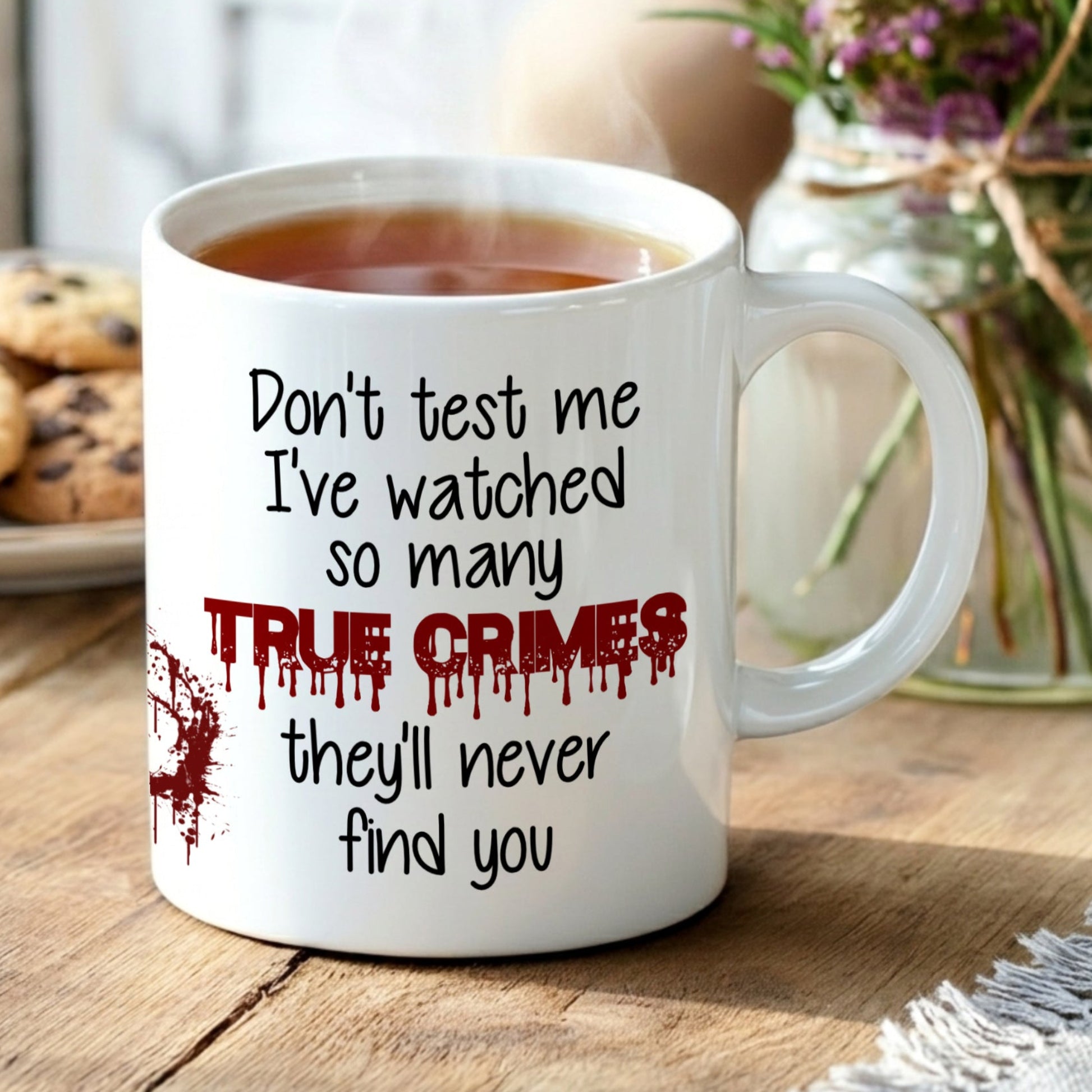 Mug with text 'Don't test me, I've watched so many true crimes, they'll never find you' on a wooden table with cookies and flowers.