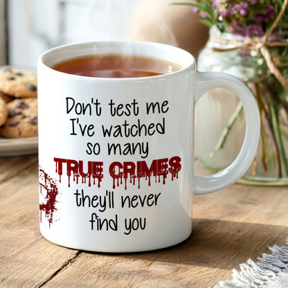 Mug with text 'Don't test me, I've watched so many true crimes, they'll never find you' on a wooden table with cookies and flowers.