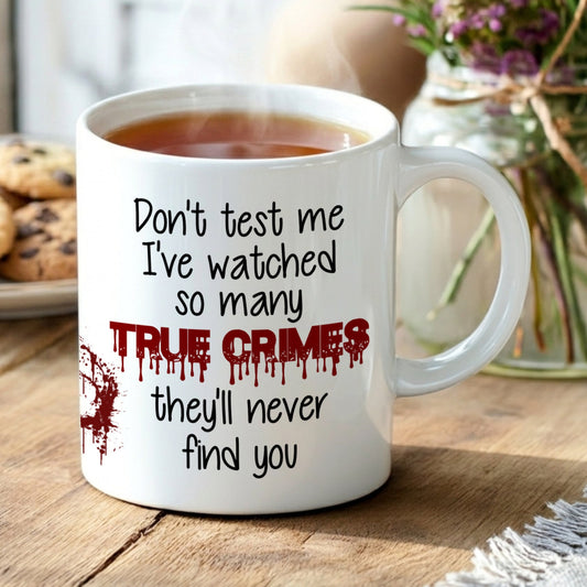 Mug with text 'Don't test me, I've watched so many true crimes, they'll never find you' on a wooden table with cookies and flowers.
