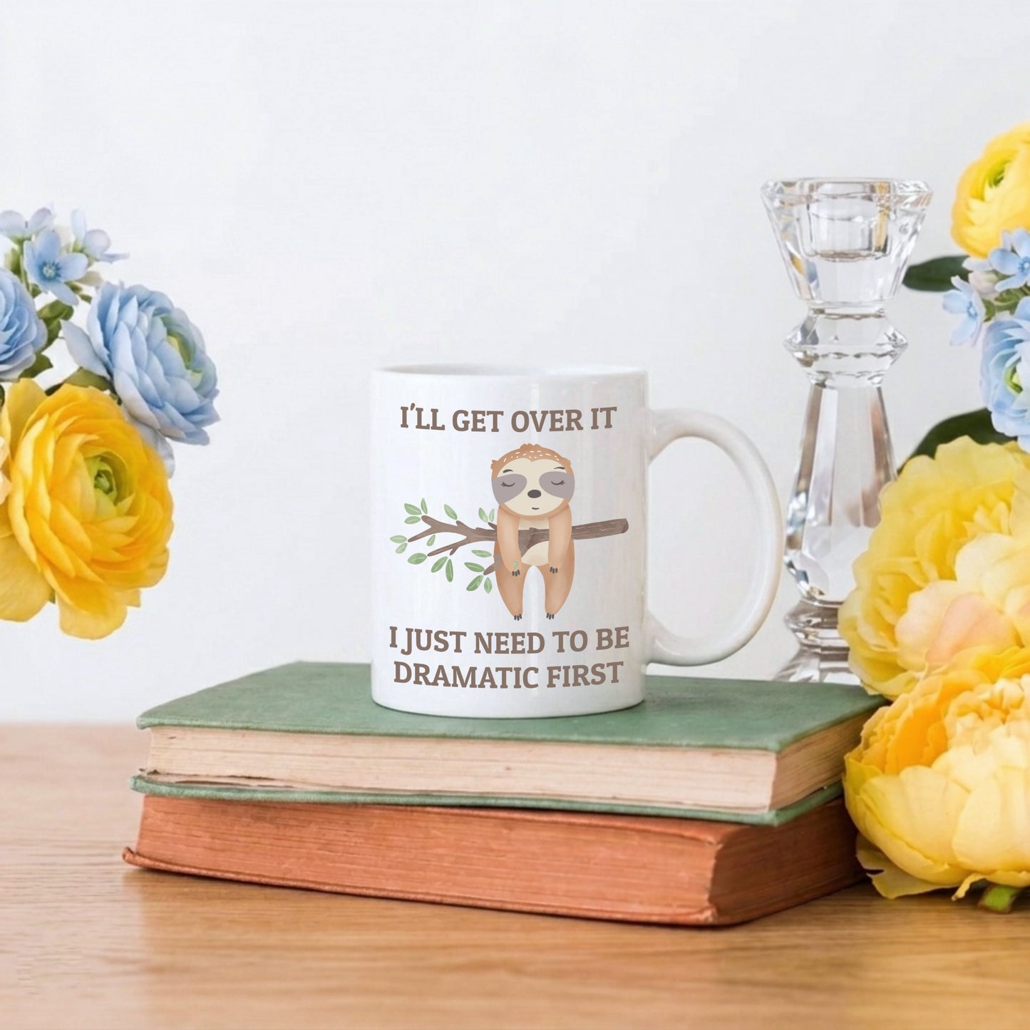 Mug with sloth graphic and text on a stack of books with flowers and a candle in the background