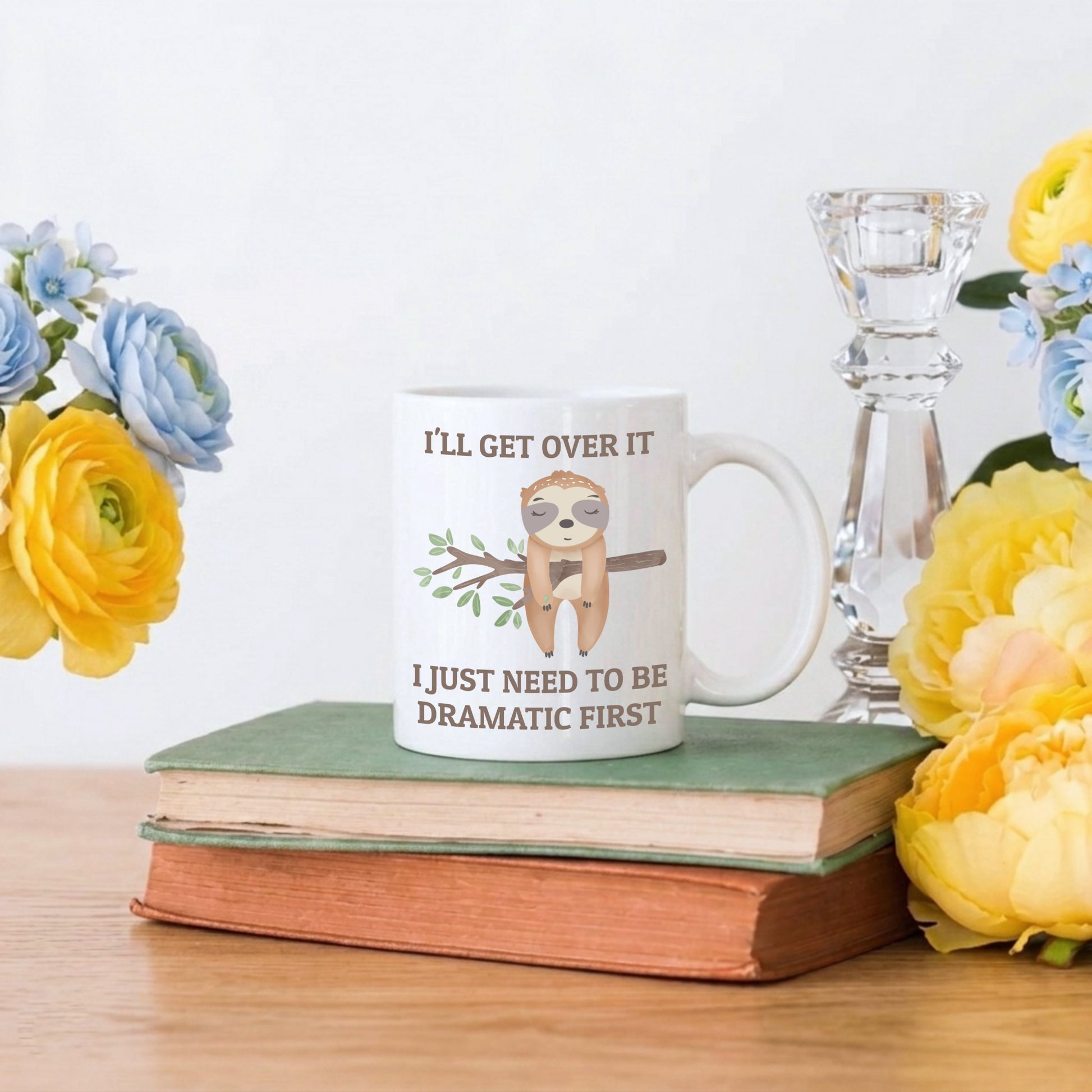Mug with sloth graphic and text on a stack of books with flowers and a candle in the background