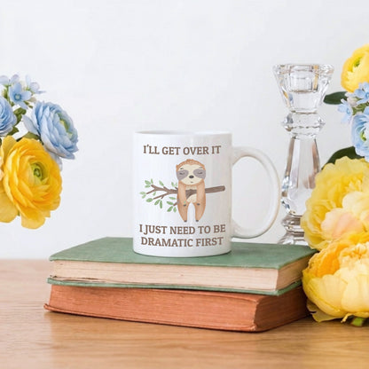 Mug with sloth graphic and text on a stack of books with flowers and a candle in the background