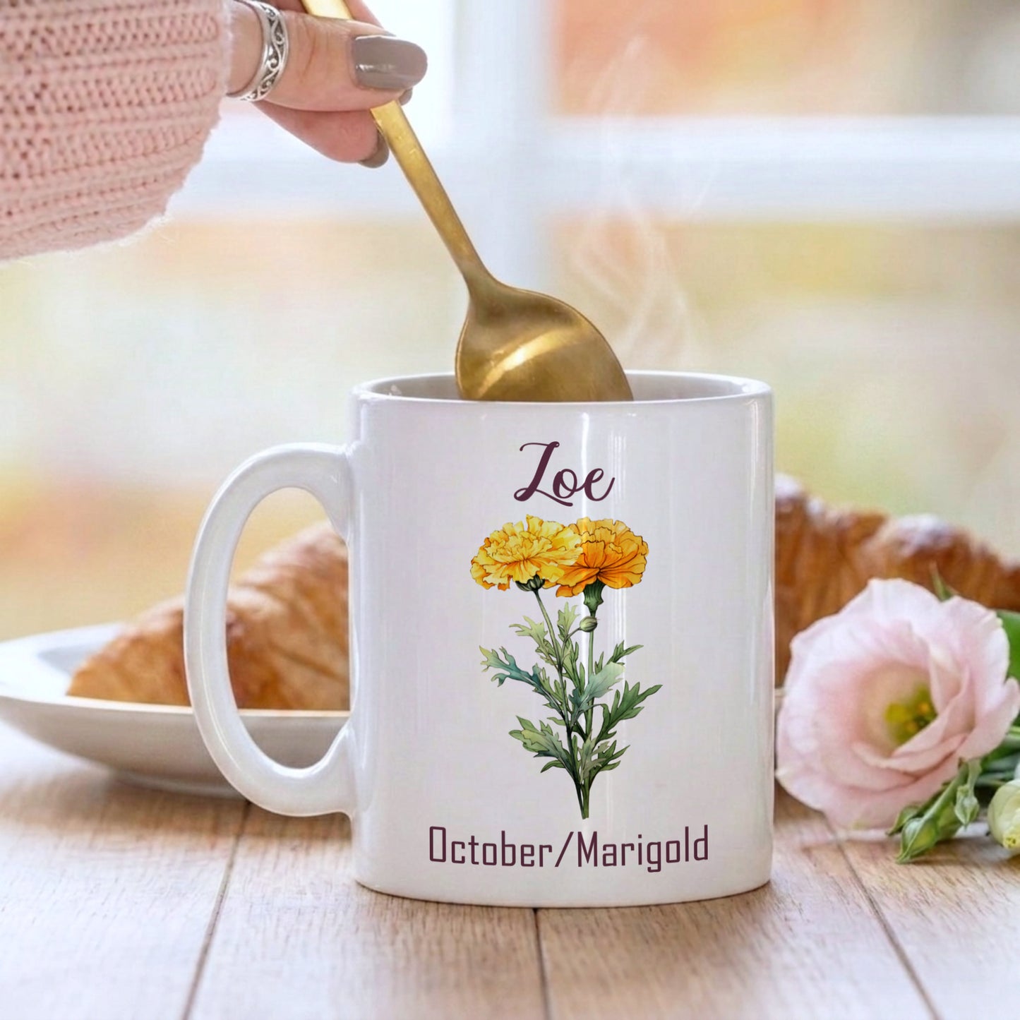 White mug with floral design and 'Loe' text, held by a hand with a gold spoon, on a wooden surface with a croissant and flowers.