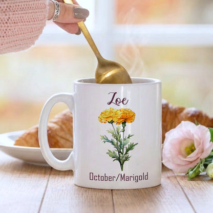 White mug with floral design and 'Loe' text, held by a hand with a gold spoon, on a wooden surface with a croissant and flowers.