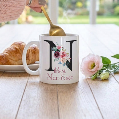 A white ceramic mug with a large black letter N, floral accents, and the words 'Best Nan Ever' in pink script, set on a wooden table in a bright room. A hand in a pink sweater stirs a gold spoon into the mug, with a croissant on a plate and a pink flower nearby.