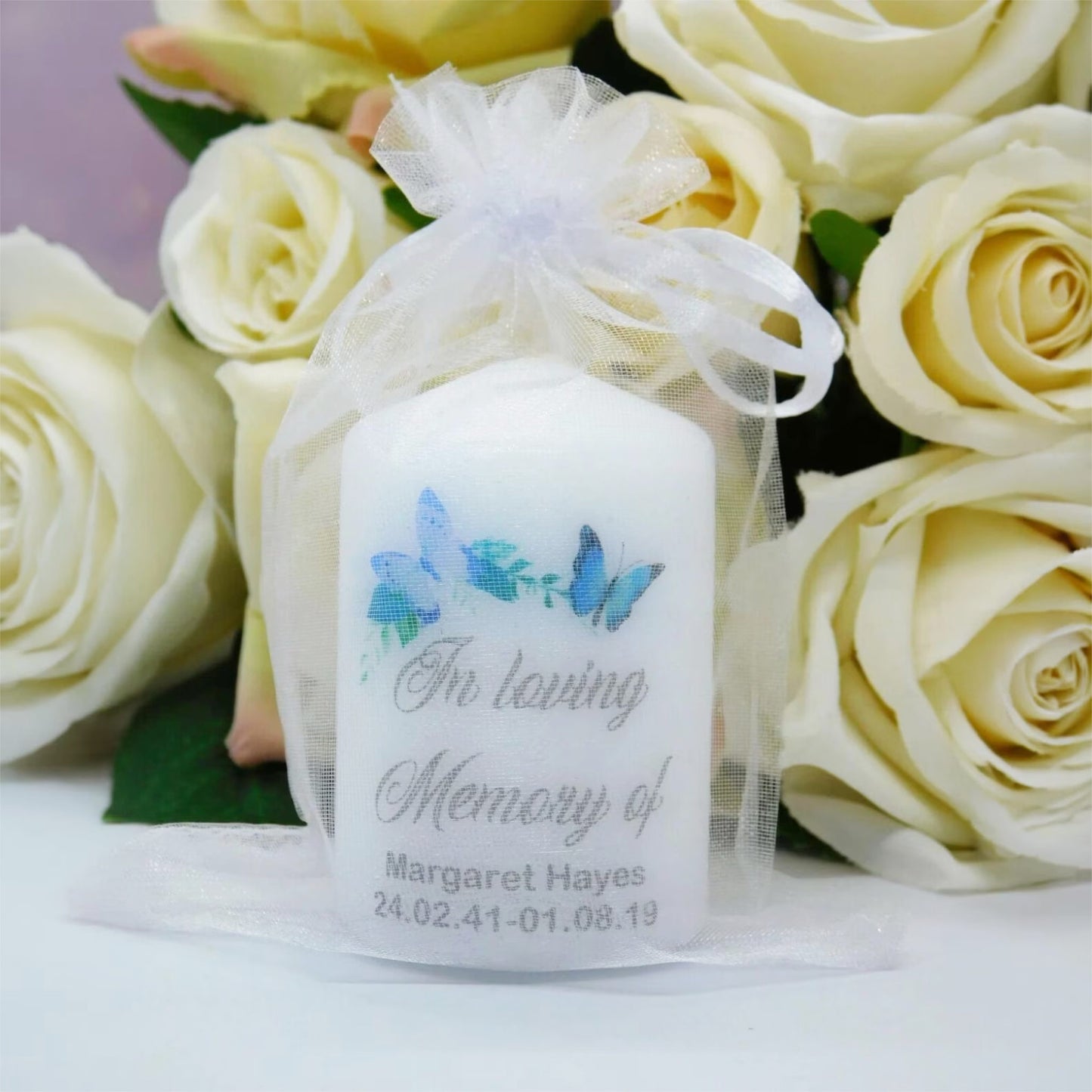 A personalised white memorial candle in a sheer white organza bag, decorated with blue butterflies and the text 'In loving Memory of Margaret Hayes' with a date. The candle is styled in front of a bouquet of pale yellow roses.