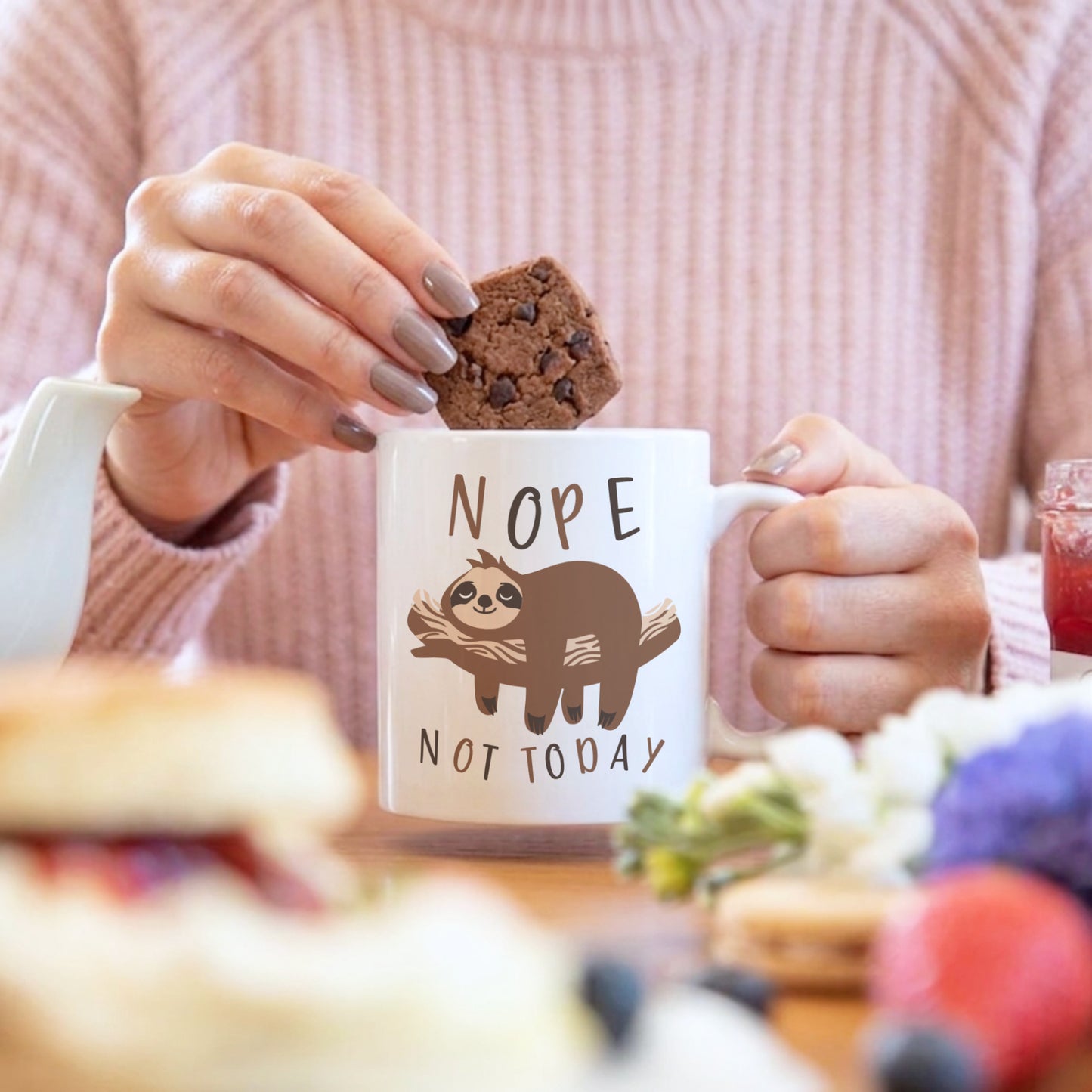 'Nope Not Today' sloth mug held by a woman dunking a chocolate brownie, with a fruit drink and baked goods in the background
