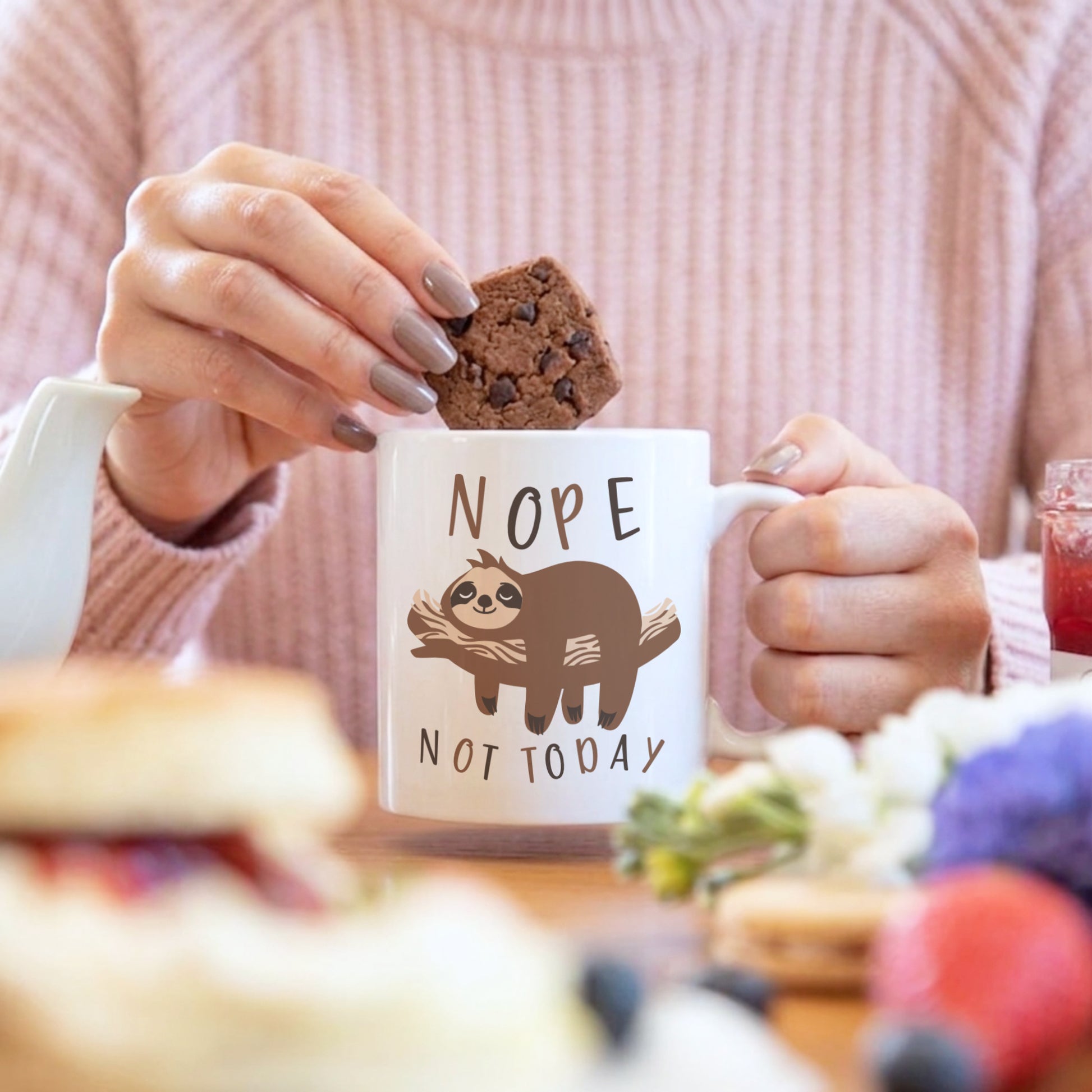 'Nope Not Today' sloth mug held by a woman dunking a chocolate brownie, with a fruit drink and baked goods in the background