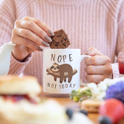 'Nope Not Today' sloth mug held by a woman dunking a chocolate brownie, with a fruit drink and baked goods in the background