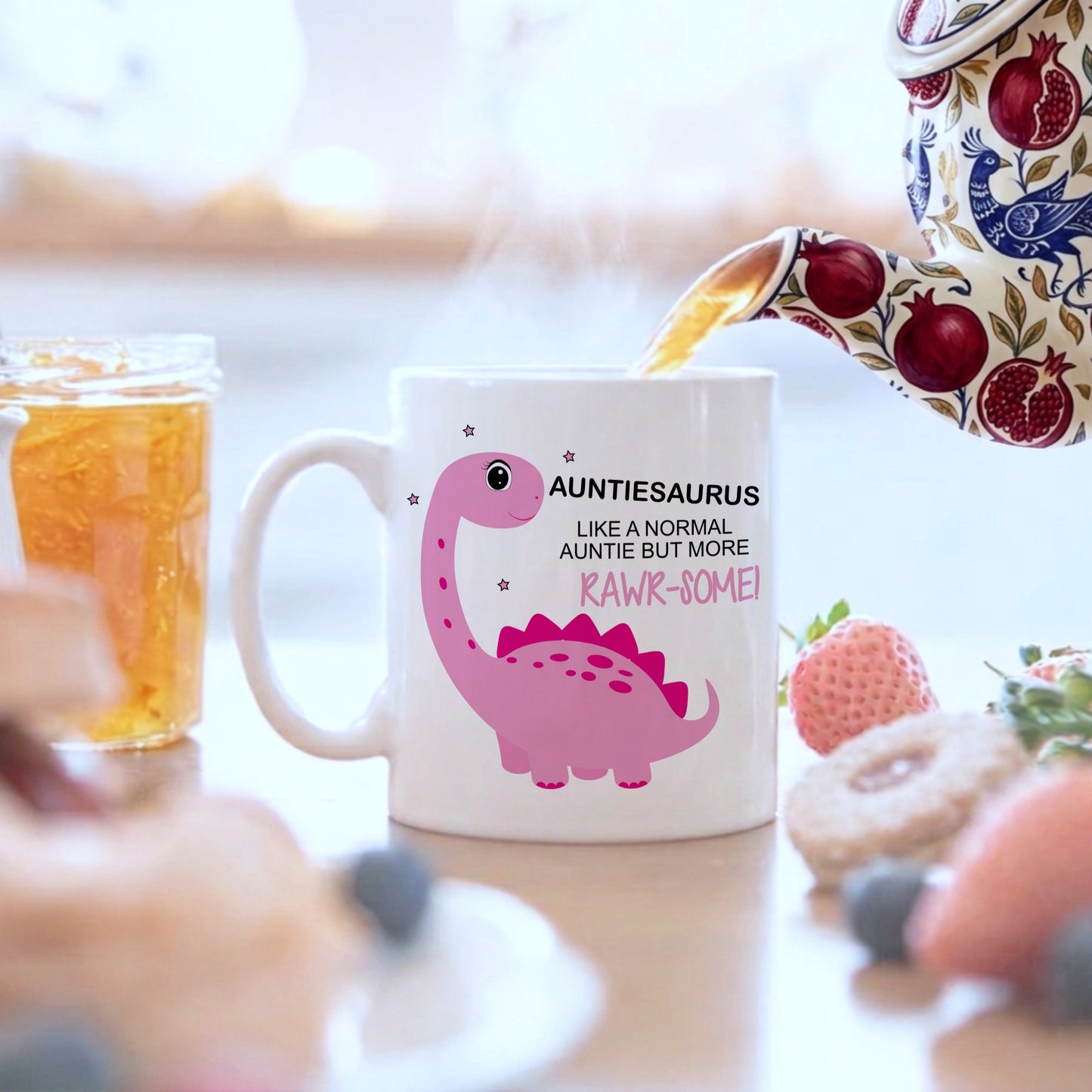 A white ceramic mug with a pink dinosaur graphic and the text 'AUNTIESAURUS LIKE A NORMAL AUNTIE BUT MORE RAWK-SOME!' sits on a table with a teapot pouring tea into it, surrounded by breakfast items like a strawberry, blueberries, and a honey jar.