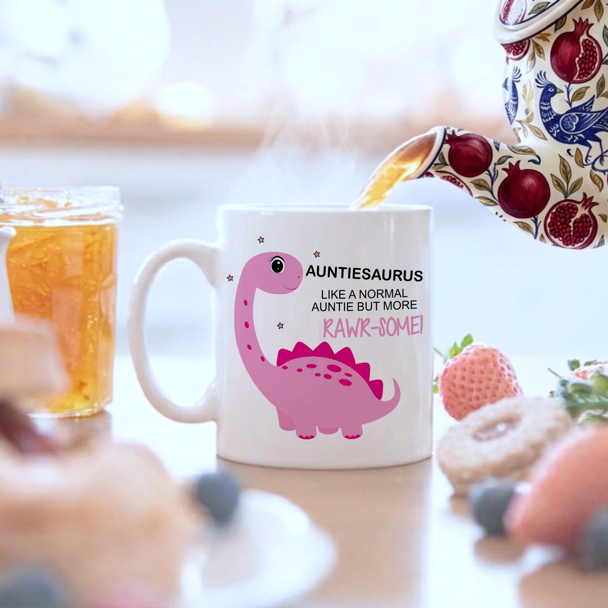 A white ceramic mug with a pink dinosaur graphic and the text 'AUNTIESAURUS LIKE A NORMAL AUNTIE BUT MORE RAWK-SOME!' sits on a table with a teapot pouring tea into it, surrounded by breakfast items like a strawberry, blueberries, and a honey jar.