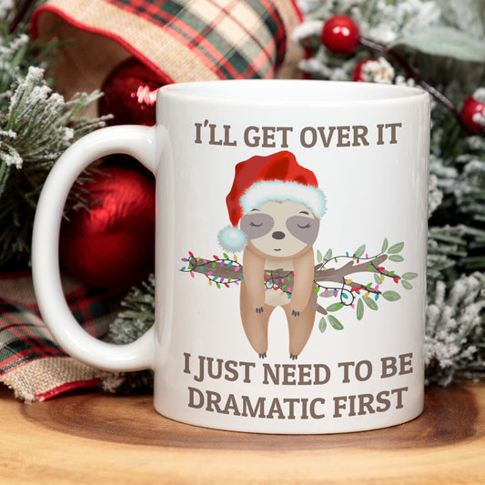 White mug with sloth illustration and Christmas-themed text, surrounded by festive decorations.