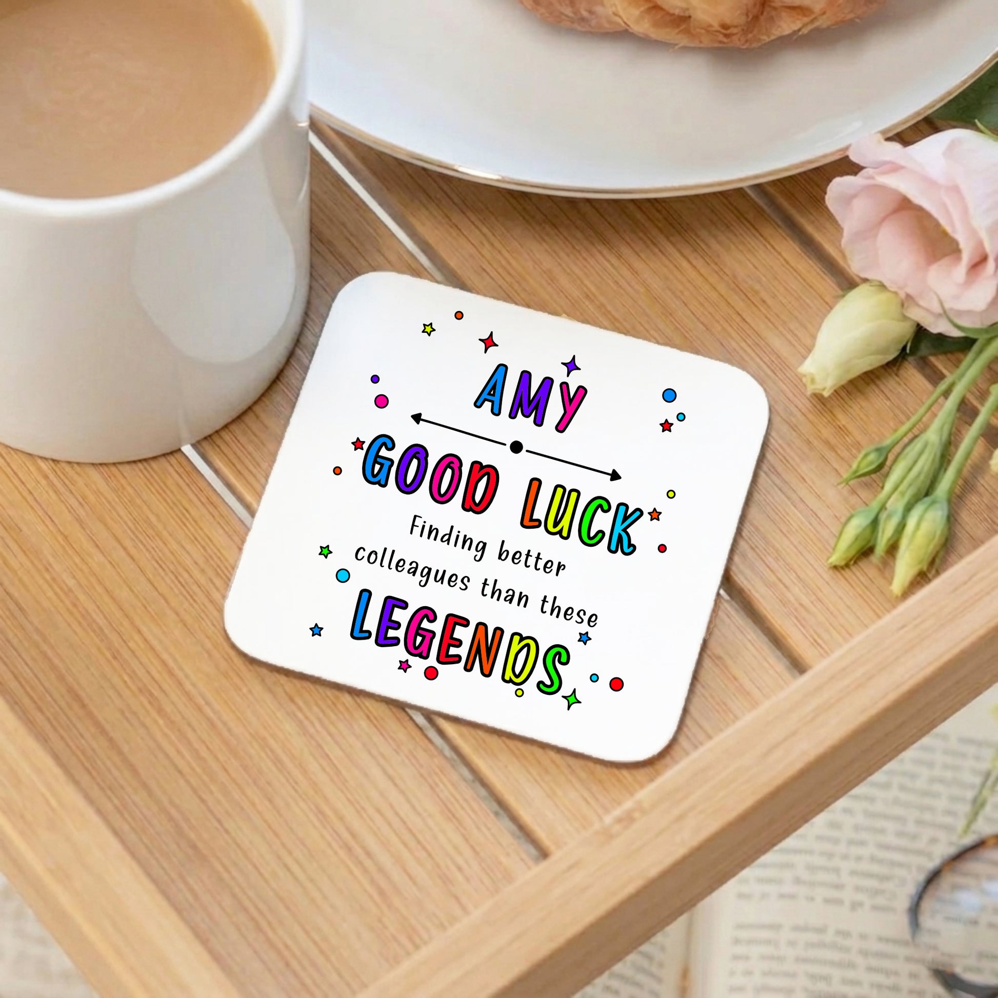 Coaster with colourful text on a wooden table with a mug and flowers.