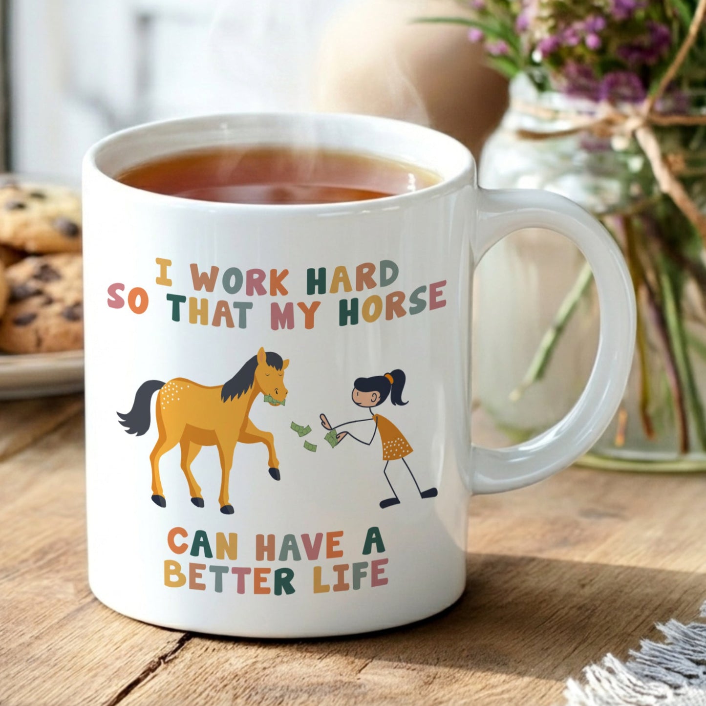 Mug with horse and person illustration and text on a wooden table with tea and cookies.