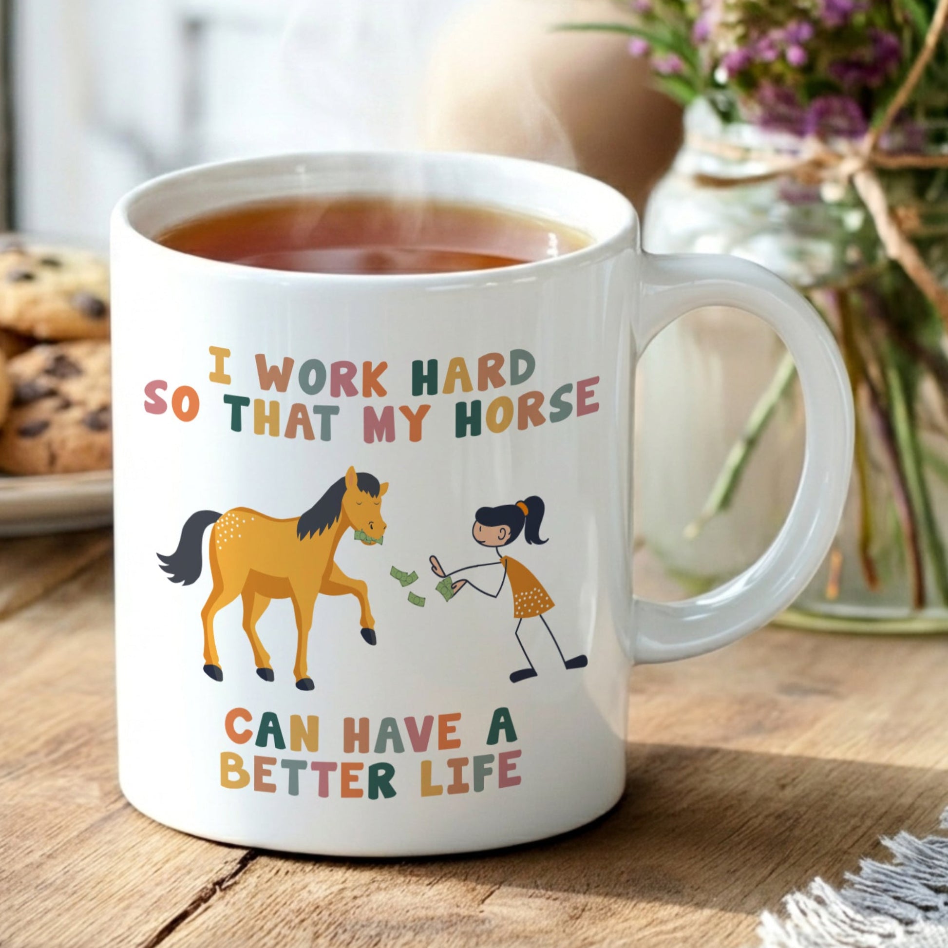 Mug with horse and person illustration and text on a wooden table with tea and cookies.