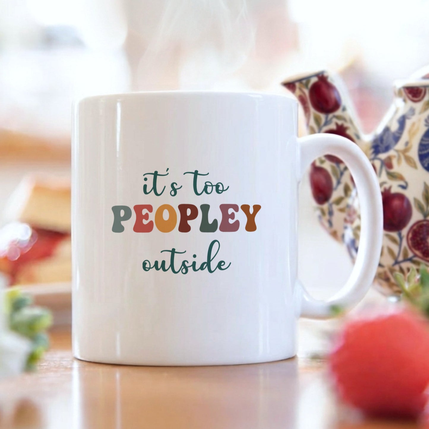 White mug with colorful text 'it's too peopley outside' on a blurred background