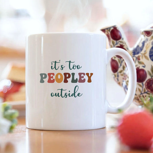 White mug with colorful text 'it's too peopley outside' on a blurred background