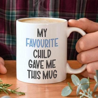 Person holding a mug with 'My favourite child gave me this mug' text on a wooden table with plants.