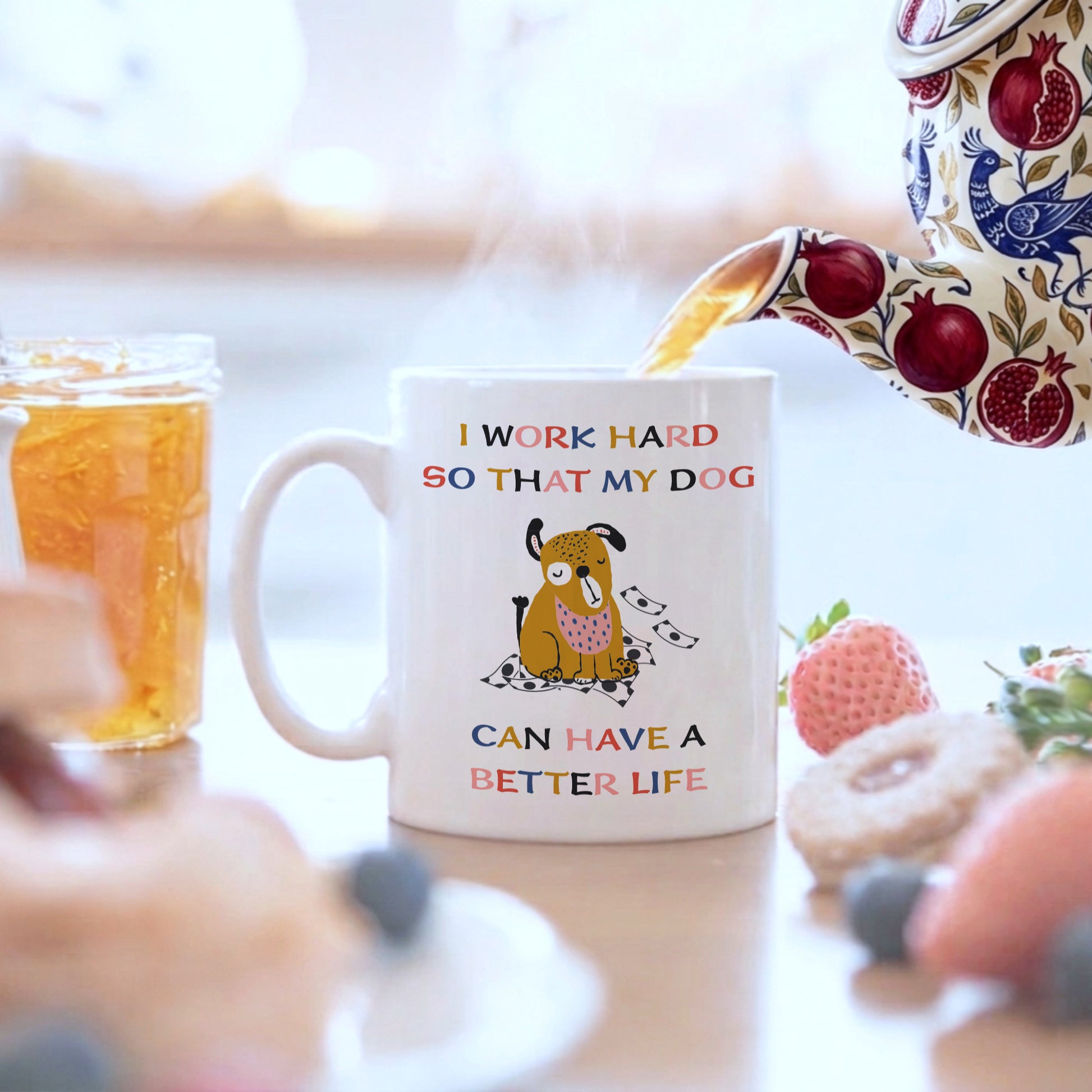 White ceramic dog lover mug with colourful 'I Work Hard So That My Dog Can Have a Better Life' quote and illustrated dog, with tea being poured from a floral teapot surrounded by fresh fruit