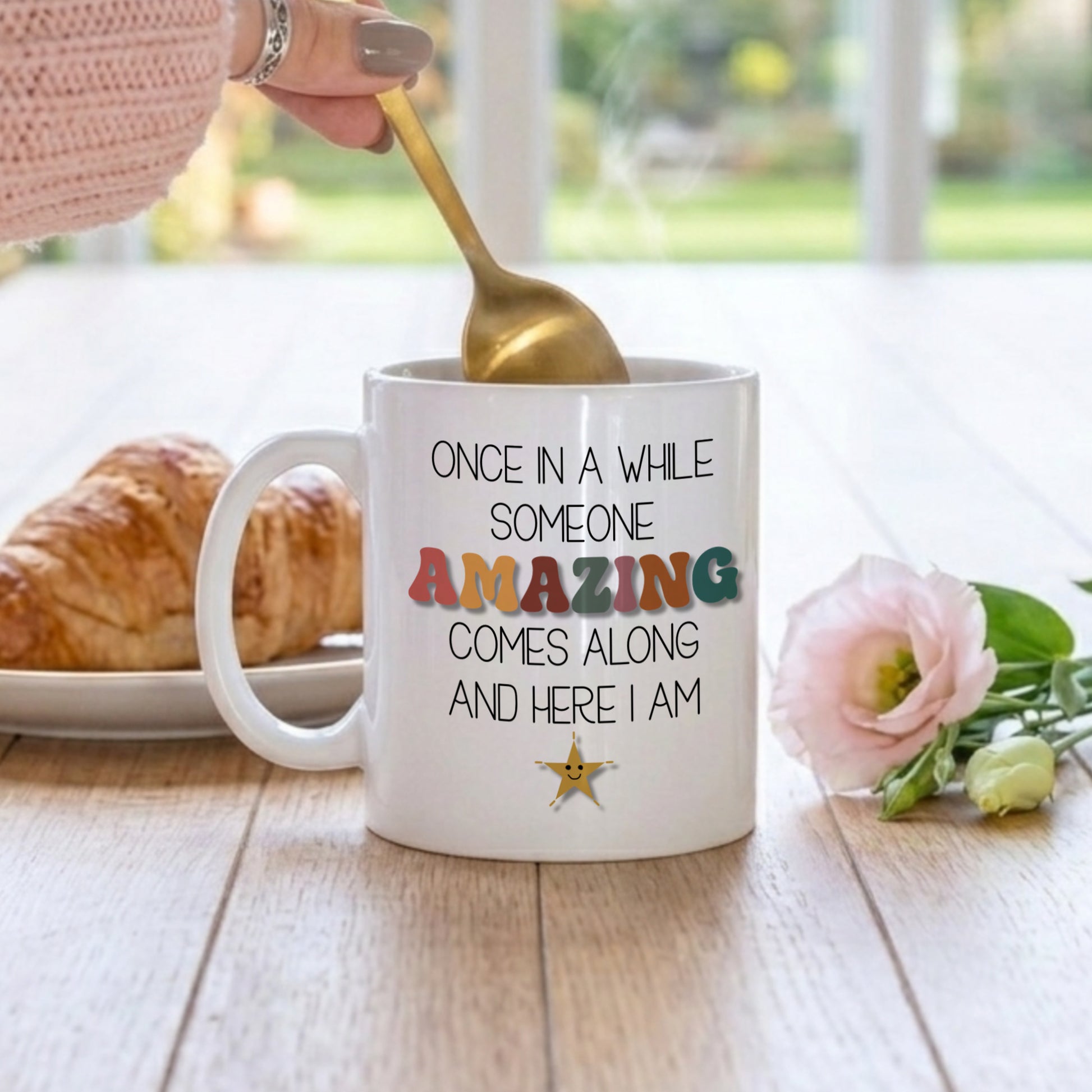 Mug with motivational quote, spoon, croissant, and flowers on a table