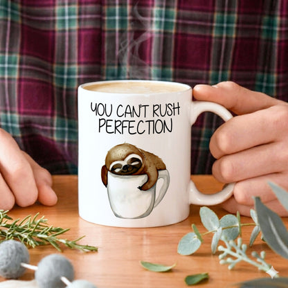 Person holding a mug with a sloth graphic and text 'You can't rush perfection' on a wooden table.
