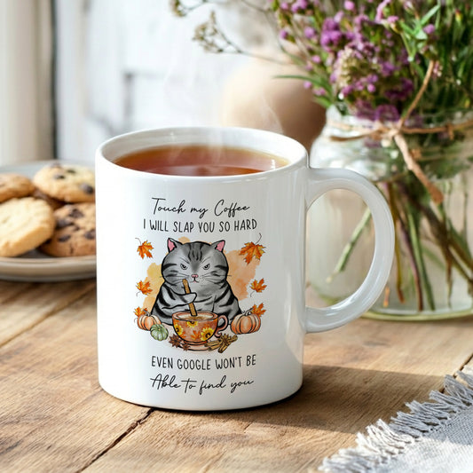 White ceramic coffee mug with a humorous cat design. The mug features a grumpy grey tabby cat with a coffee cup, surrounded by autumn leaves and pumpkins. Text on the mug reads "Touch my Coffee I WILL SLAP YOU SO HARD, EVEN GOOGLE WON'T BE Able to find you."