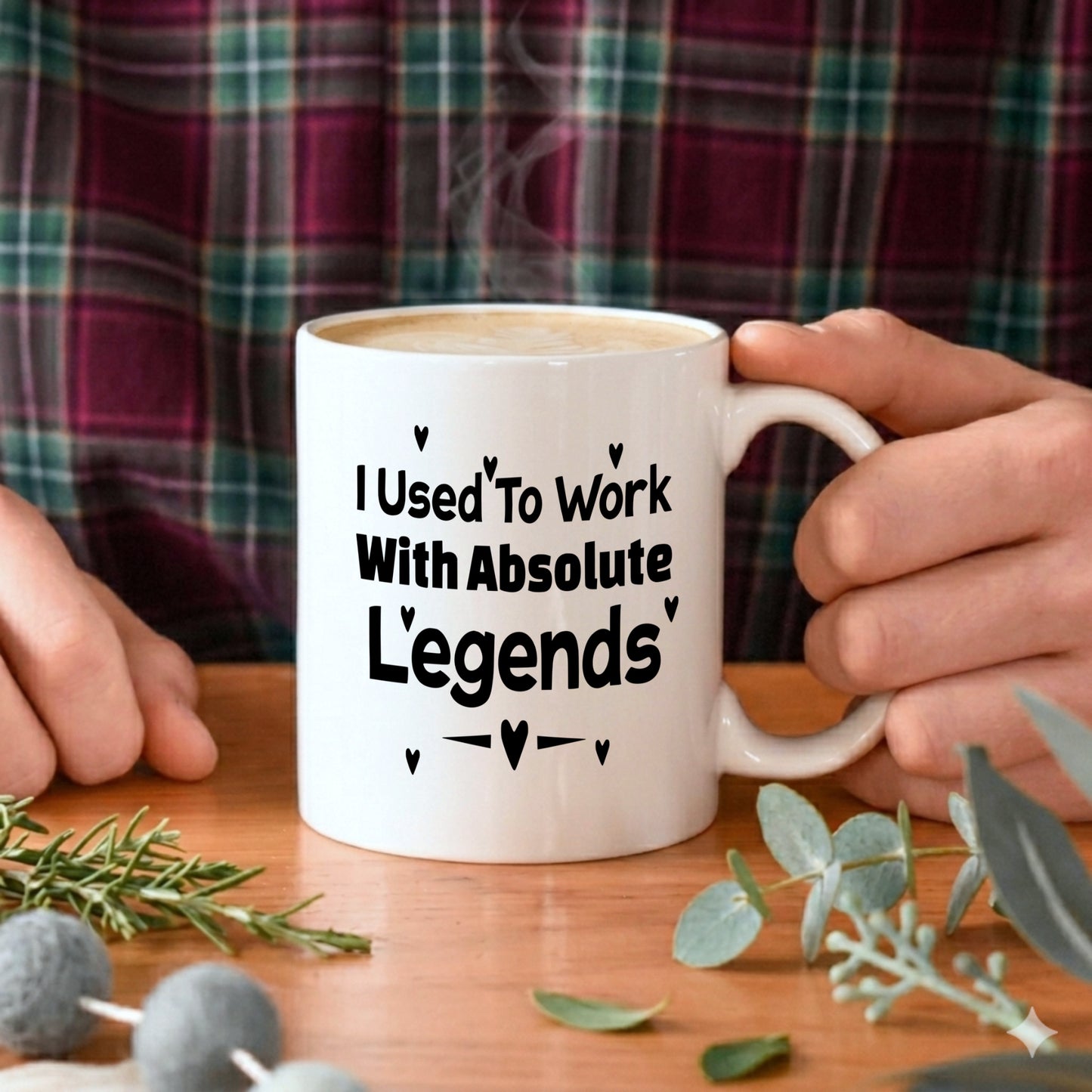 I Used To Work With Absolute Legends' white ceramic mug filled with a latte, held with both hands by a person wearing a checked shirt, with eucalyptus and foliage on the table