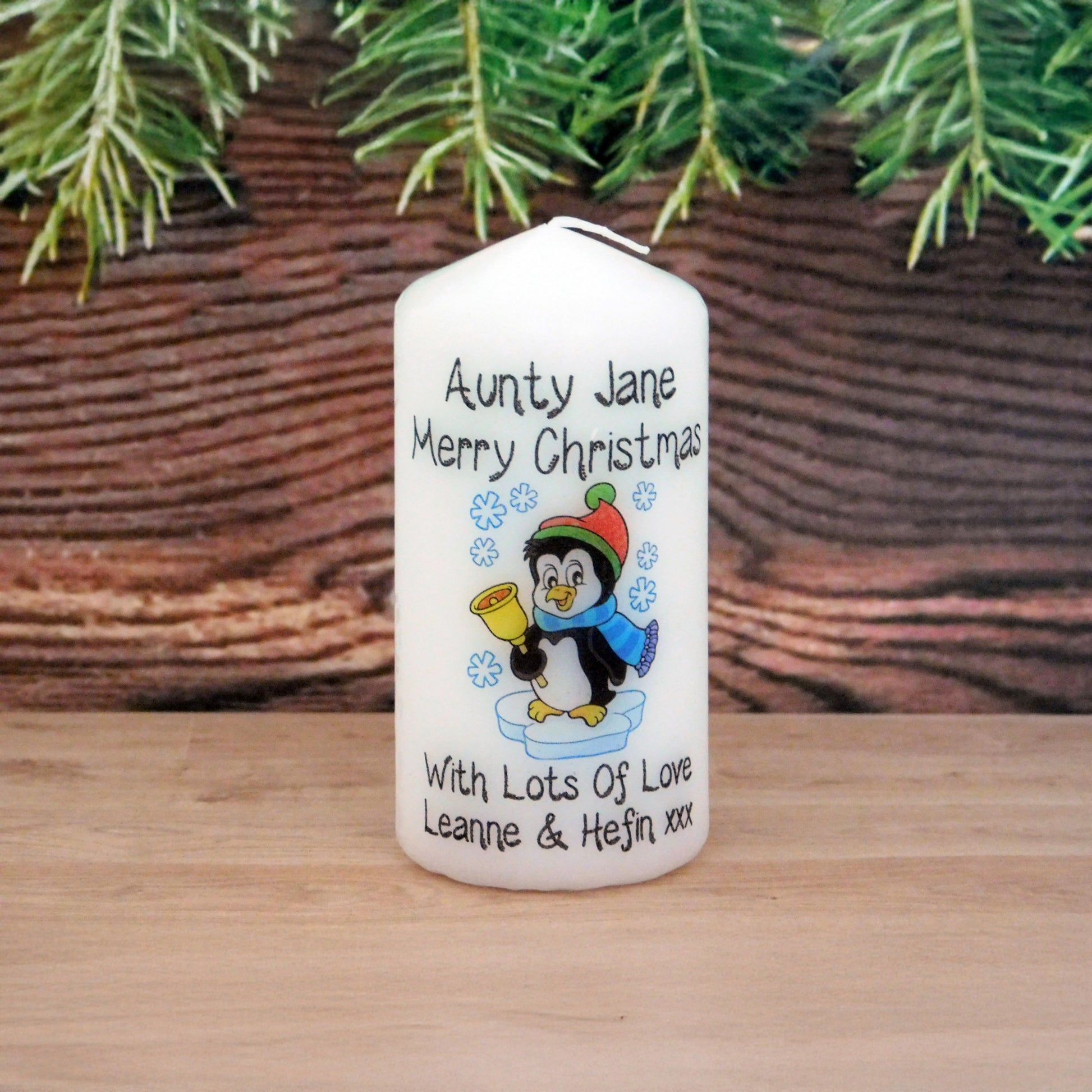 Personalized Christmas candle with a penguin design and text, placed against a wooden background with greenery.