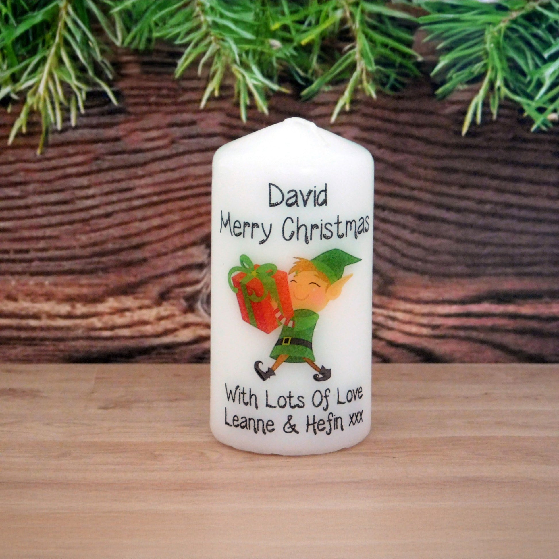 White candle with Christmas design and text on a wooden surface with greenery in the background