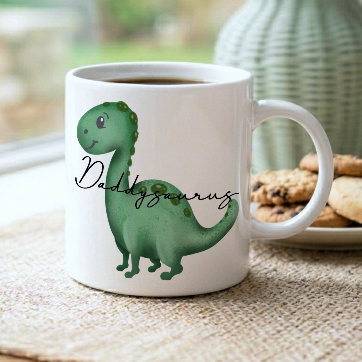 A white ceramic mug filled with coffee sits on a woven placemat in a kitchen setting, featuring a green cartoon dinosaur and the text 'Daddy Dinosaur' in black script. A plate of cookies and a green textured vase are blurred in the background.