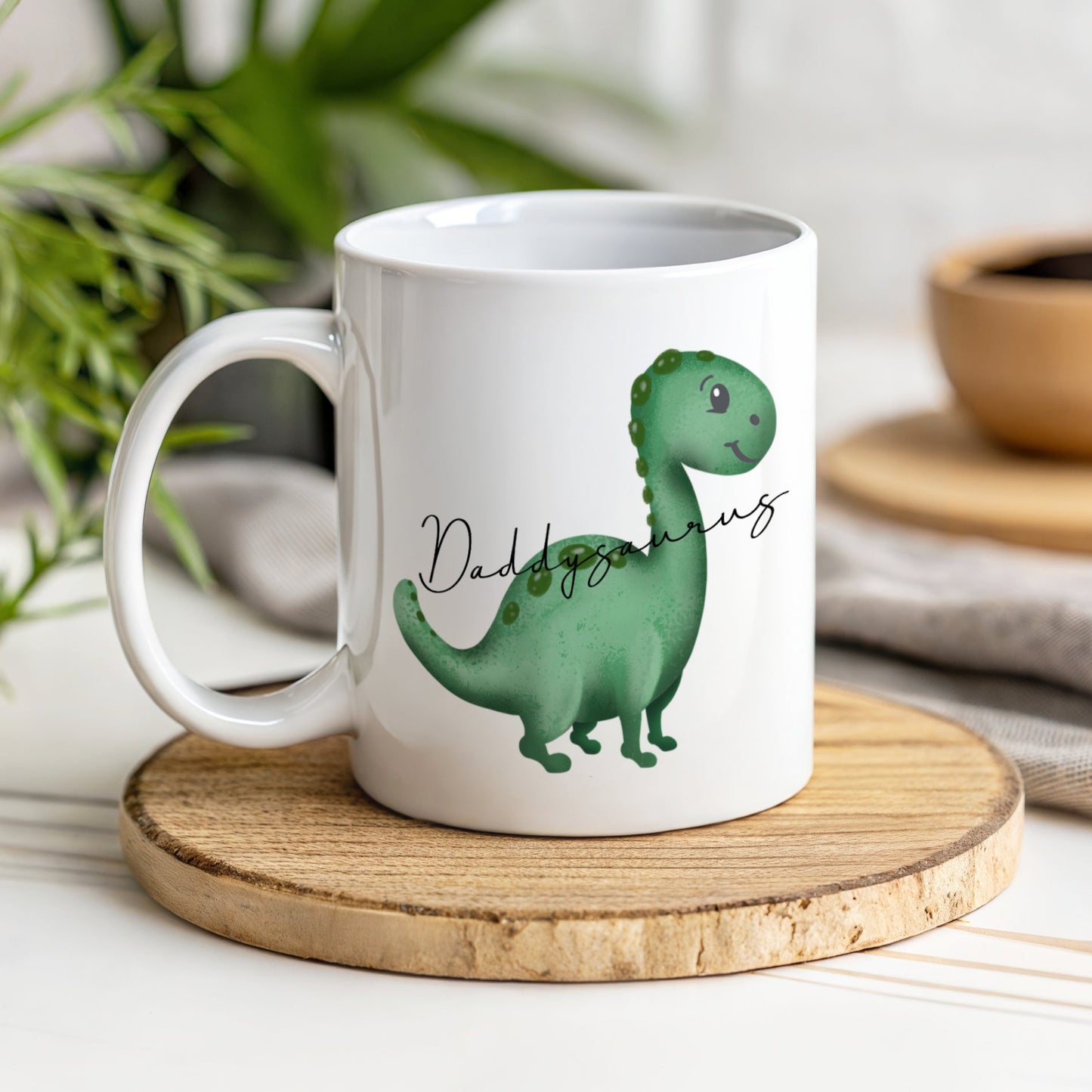 A white mug with a green dinosaur with the text Daddysaurus though the image of the dinosaur