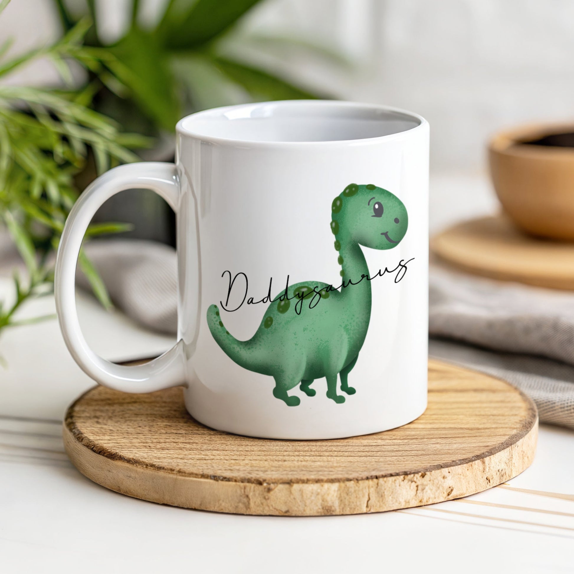 A white mug with a green dinosaur with the text Daddysaurus though the image of the dinosaur