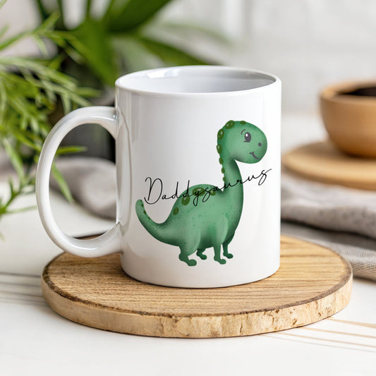 A white mug with a green dinosaur with the text Daddysaurus though the image of the dinosaur