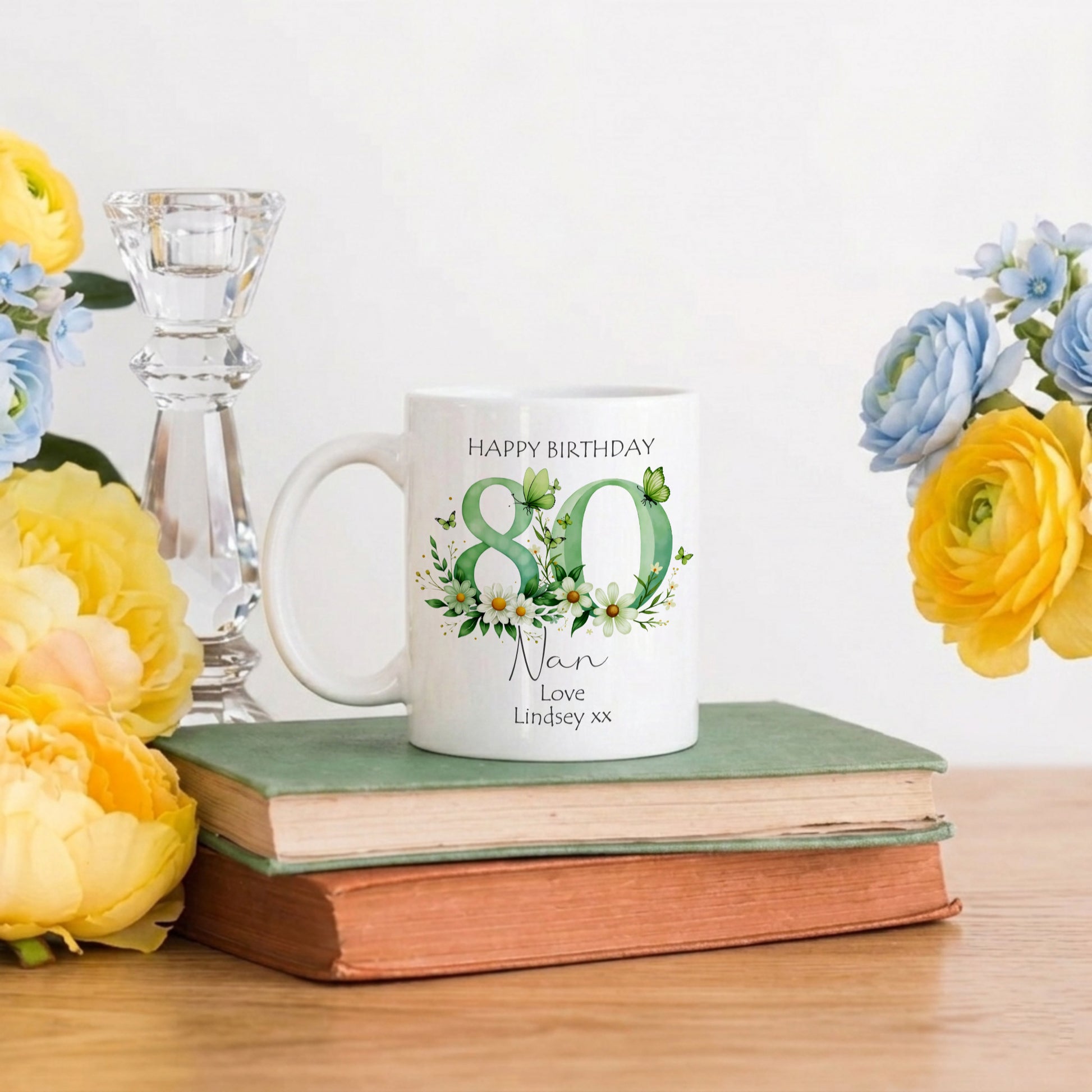 White mug with 'Happy Birthday 80 Nan' text on a stack of books with flowers around
