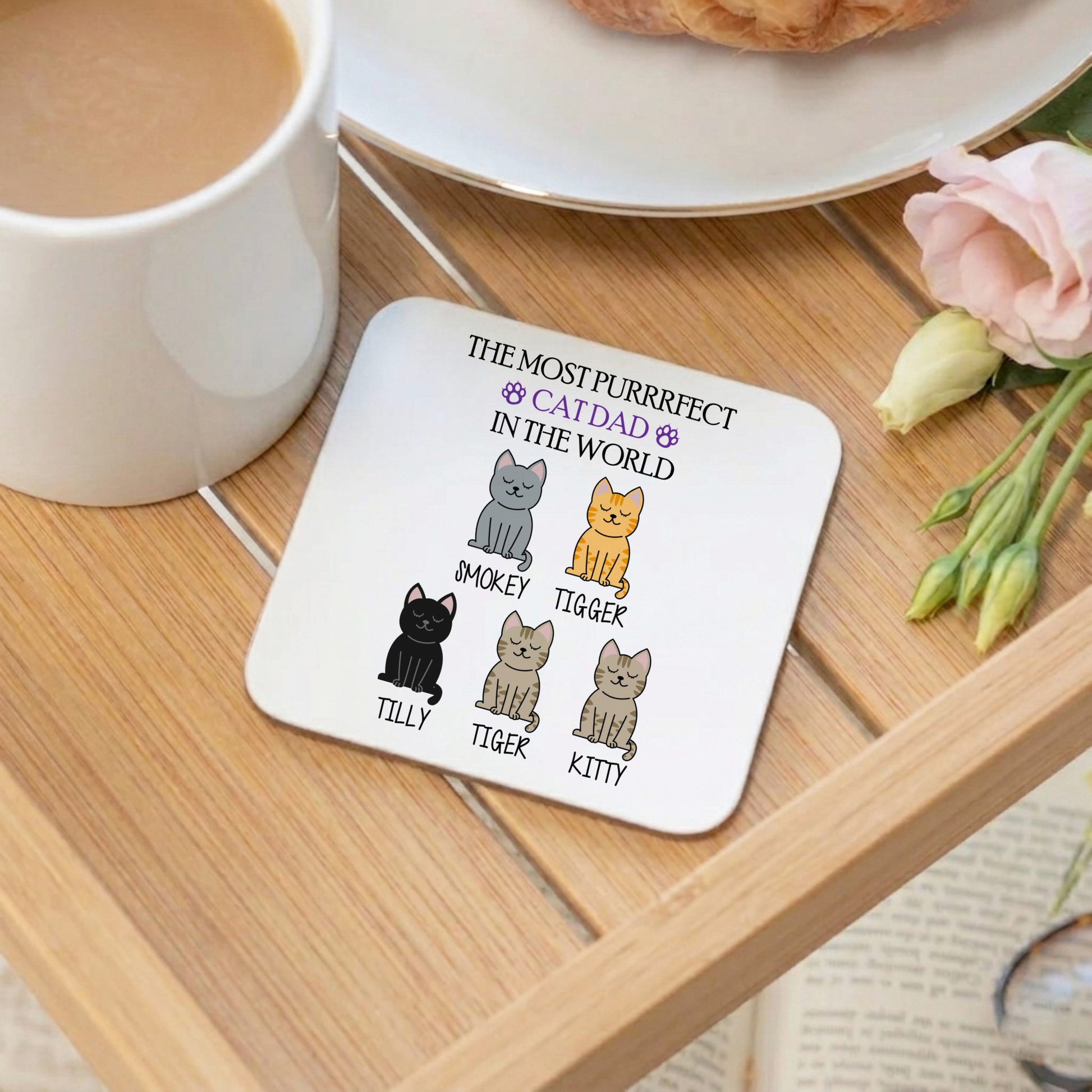 Coaster with cat illustrations and text on a wooden table with a cup of coffee and flowers.