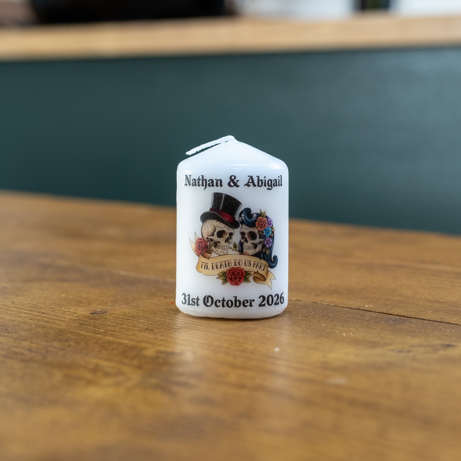 A small white personalised candle with a house-shaped top sits on a wooden table. The front of the candle is printed with black text reading 'Nathan & Abigail' and '31st October 2026' around a central illustration of two skulls wearing top hats and roses.